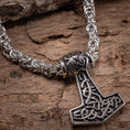 Load image into Gallery viewer, Et detaljeret nærbillede af et metallisk Mjolnir-vedhæng i form af Thors hammer, med indviklede nordiske designs, fastgjort til en robust og udsmykket flettet Halskæde Kongekæde Stål 6mm. Det vikinge-inspirerede vedhæng og kæde er lavet af rustfrit stål og vist på en robust træoverflade.
