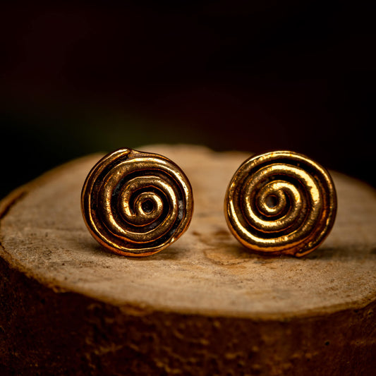 Et par Ørestikker Spiral Bronze øreringe sidder på en træoverflade mod en mørk baggrund. De polerede spiraler skinner varmt, forstærket af den subtile belysning. Det rustikke træ og de dybe skygger tilføjer en naturlig, jordnær stemning til billedet.