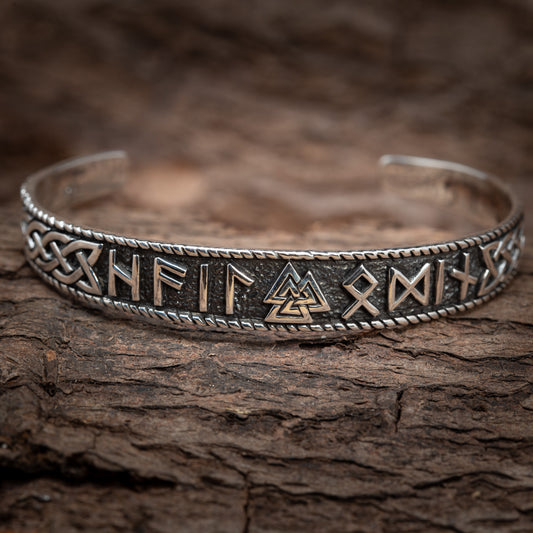 Et nærbillede af Armbånd Bangle Trial Rune 925s Sterling Sølv prydet med indviklede runedesigns og symboler, liggende på en tekstureret træoverflade. Armbåndet har udsmykkede keltiske knuder langs dets kanter og et centralt design, der ligner et sammenlåsende trekantmotiv, der minder om vikingetiden smykker.