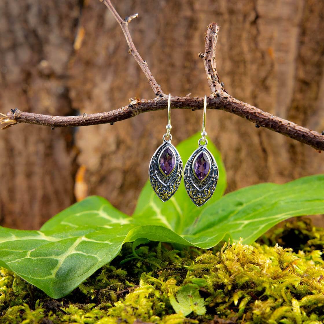 Et par elegante Hængeøreringe Braid 925s Sølv med indviklede designs og cabochon zirkon ametyst ædelsten hænger fra en rustik trægren. Under dem skaber store grønne blade og mos et levende bunddække, der forstærker de naturlige omgivelser med en skovstemning.