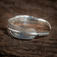 Load image into Gallery viewer, "Armbånd Bangle Feather 925s Sterling Sølv" er et 925s sterlingsølv armbånd, der er indviklet designet til at ligne en detaljeret kragefjer, med den ene ende snoet rundt for at danne det åbne armbånd, smukt hvilende på en tekstureret træoverflade.
