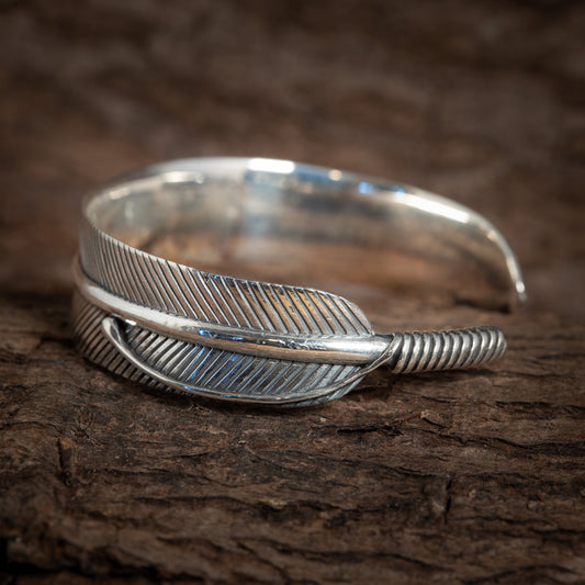 "Armbånd Bangle Feather 925s Sterling Sølv" er et 925s sterlingsølv armbånd, der er indviklet designet til at ligne en detaljeret kragefjer, med den ene ende snoet rundt for at danne det åbne armbånd, smukt hvilende på en tekstureret træoverflade.