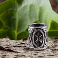 Load image into Gallery viewer, Et nærbillede af en detaljeret Vikingesmykker-ring med indviklede mønstre og et centralt "X"-design, der minder om Rune Gebo. Skægperler / Hårpynt Rune G Gebo 925s Sølv ringen er placeret på en struktureret, barklignende overflade med et stort grønt blad i baggrunden.
