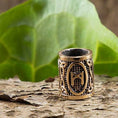 Load image into Gallery viewer, En detaljeret, cylindrisk ring med en graveret Mannaz-rune, kendt som Skægperler / Hårpynt Rune M Bronze, hviler på en struktureret, bark-lignende overflade. Baggrunden viser store, levende grønne blade. Ringen ligner vikingesmykker og ser ud til at være lavet af et metallisk materiale, muligvis bronze.
