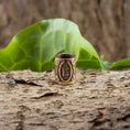 Load image into Gallery viewer, Skægperler / Hårpynt Rune P Pertho Bronze, et indviklet udskåret bronzestykke, der minder om vikingesmykker, vises på en struktureret barkoverflade med et grønt blad i baggrunden, der giver en naturlig, jordnær indstilling.
