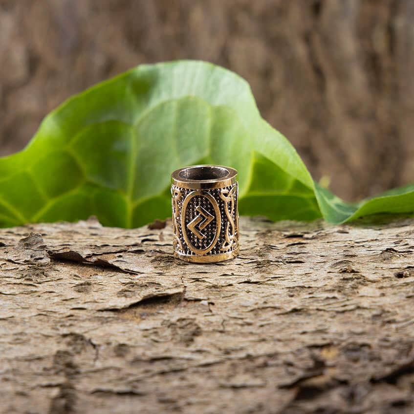 En omhyggeligt udformet ring, med det indviklede design af Skægperler / Hårpynt Rune J Jera Bronze, hviler yndefuldt på et stykke træbark. Et stort grønt blad i baggrunden fremhæver dets udsøgte geometriske former og delikate udskæringer, der minder om vikingesmykker, hvilket skaber en slående kontrast mod de naturlige, rustikke omgivelser.