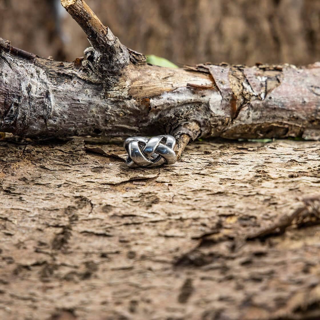 En smukt vævet Skægperler / Hårpynt Vævet Lille Stål hviler på en barsk træstamme, der blander sig problemfrit med sine naturlige omgivelser. Baggrunden viser tekstureret bark og grene, hvilket giver en jordagtig, rustik følelse, der fremhæver ringens autentiske udseende.