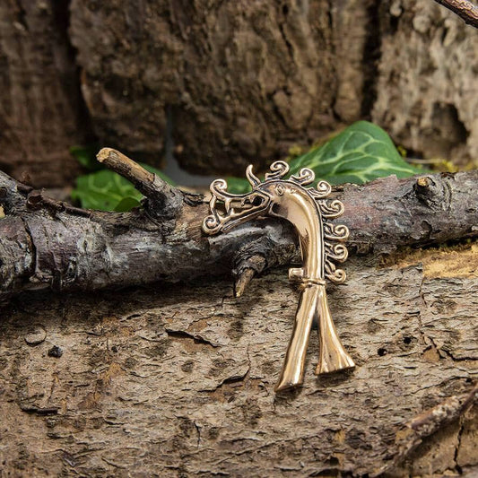 Et nærbillede viser en Broche Drage Bronze i form af en drage, der hviler på ru bark. Brochen har indviklede detaljer med hvirvlende mønstre, der fanger essensen af en mytisk drage. Grønne blade i baggrunden tilføjer et strejf af natur, der minder om Birka Sveriges ældgamle tiltrækning.