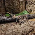 Load image into Gallery viewer, En Broche Urnesstil Bronze hviler på et stykke forvitret træ, omgivet af tekstureret bark og grønne blade i baggrunden. Lys giver subtil vægt på de indviklede detaljer i dets autentiske bronzemønster.
