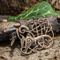 Load image into Gallery viewer, Broche Urnesstil Bronze, prydet med indviklede keltiske knudedesigns med stiliserede dyrehoveder, hviler på et stykke træbark. Denne autentiske bronzebroche fremkalder ånden fra Tröllaskógur-øen med grønne blade i baggrunden, der forstærker dens tidløse skønhed.
