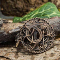 Load image into Gallery viewer, Broche Pitney Spænde Bronze, en autentisk bronze museumskopi, viser indviklede keltisk-inspirerede designs med detaljerede sammenvævede mønstre. Afbildet mod ru træbark og grønne blade i baggrunden har brochen en blanding af løkker og knuder inden for en cirkulær ramme, der minder om Urnestil-perioden.
