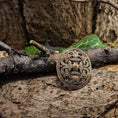 Load image into Gallery viewer, En bronze Broche Borrestil Bronze hviler på en barkdækket skovbund omgivet af grene og grønt løv. Brochen har indviklede, sammenflettede mønstre, der afspejler gammelt håndværk. Scenen fremkalder en følelse af historie og naturens omfavnelse, der minder om autentisk vikingedesign.
