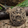 Load image into Gallery viewer, Broche Borrestil Bronze, en kompliceret designet broche i middelalderstil, der minder om autentisk vikingedesign, hviler på et stykke forvitret træ. Brochen har et udsmykket, symmetrisk mønster med sammenflettede detaljer og en central hævet knude, sat mod en rustik baggrund med grønt løv, der fanger essensen af Fæsted skatten.
