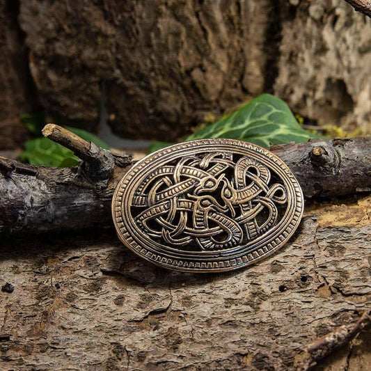 "Broche Jellingstil Bronze", en oval broche, der er indviklet designet med sammenflettede knaster og symboler, der minder om Jellinge-stilen, hviler på et groft stykke træ omgivet af grønne blade. Denne autentiske bronzebroche har et gammelt, antikt udseende, der antyder dens historiske eller kulturelle betydning fra vikingetiden.