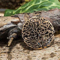 Load image into Gallery viewer, "Broche Yggdrasil Livets Træ Bronze", et indviklet udskåret rund bronzevedhæng, vises på en tekstureret træoverflade. Grøn vegetation i baggrunden tilføjer et naturligt præg til scenen.
