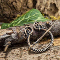 Load image into Gallery viewer, Et nærbillede af Broche Ringnål Antik Bronze, med indviklede løkker og et snoet design, der minder om traditionelle vikingebrocher, placeret på ru bark. I baggrunden skaber grønne blade og slørede naturlige elementer en udendørs rustik ramme.
