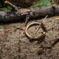 Load image into Gallery viewer, Broche Ringnål Stor Bronze, prydet med indviklede keltiske designs, hviler på et tekstureret stykke træ. Med trægrene og grønne blade i baggrunden fremkalder denne bronzering minder om vikingegenstande med sine detaljerede graveringer, der har hjertelignende former og spiraler.
