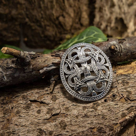 En indviklet designet Broche Borrestil 925s Sterling Sølv med keltiske knudemønstre er placeret på et tekstureret, forvitret stykke træ, ledsaget af nogle grønne blade i baggrunden, hvilket fremhæver en naturlig og rustik indstilling. Perfekt til historieinteresseret, dette stykke fanger tidløs elegance og historisk charme.