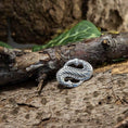 Load image into Gallery viewer, En detaljeret Broche Midgårdsormen 925s Sterling Sølv med en sammenflettet, tekstureret knude er placeret på en ru bark. Grønne blade giver en naturlig baggrund, der forstærker den rustikke og jordiske æstetik ved dette Vikingetiden-symbol.
