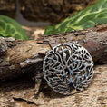 Load image into Gallery viewer, Broche Yggdrasil Livets Træ 925s Sterling Sølv, et rundt sølvvedhæng med et indviklet Yggdrasil-design, hviler på et stykke bark. Baggrunden har frodige grønne blade, der skaber en naturlig indstilling, der fremhæver vedhængets detaljerede håndværk.
