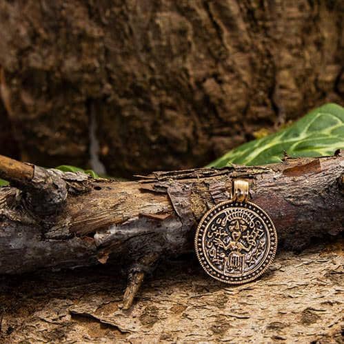 Amuletten Frigg Vedhæng Bronze, en rund medaljon med indviklede designs og symboler, hviler mod en ru, forvitret trægren på en skovbund. Baggrunden inkluderer grønne blade og træets teksturerede bark, hvilket forstærker den rustikke, naturlige scene.