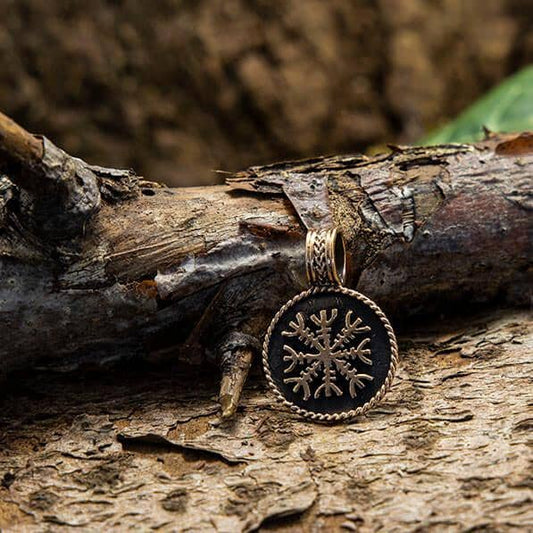 Indkapslet i en guldramme ligger Ægirshjelm Amulet Vedhæng Bronze, med et detaljeret snefnugdesign, på en tekstureret træoverflade udendørs. Baggrunden inkluderer et stykke bark og noget løv, hvilket giver bronzeamuletten et rustikt og omhyggeligt udformet udseende.