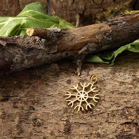 En Rune Amulet Drømme Vedhæng Forgyldt 925s Sterling Sølv vedhæng, designet som et snefnug eller indviklet hjul, ligger på en struktureret barkflade omgivet af grønne blade og en forvitret trægren. De naturlige omgivelser understreger smukt amulettens delikate håndværk.