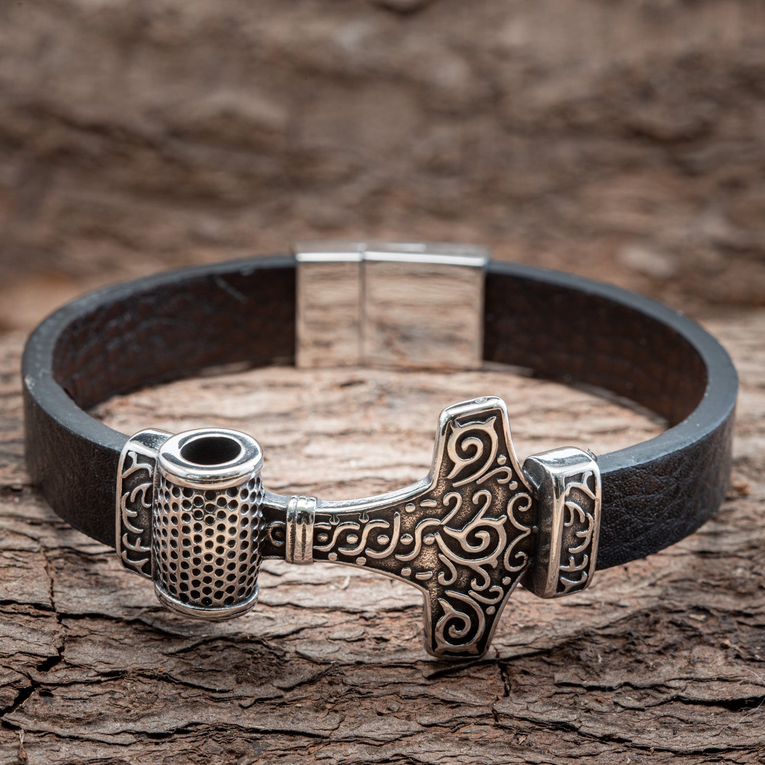 Et nærbillede af Armbånd Thorshammer m. Mønster Stål, der viser sin stilfulde sorte læderrem og en sølvfarvet Thors Hammer (Mjolnir) midtpunkt prydet med indviklede nordiske designs. Armbåndet vises på en tekstureret træbaggrund, der fremhæver dets detaljerede håndværk og suppleret med rustfrit stål-elementer.