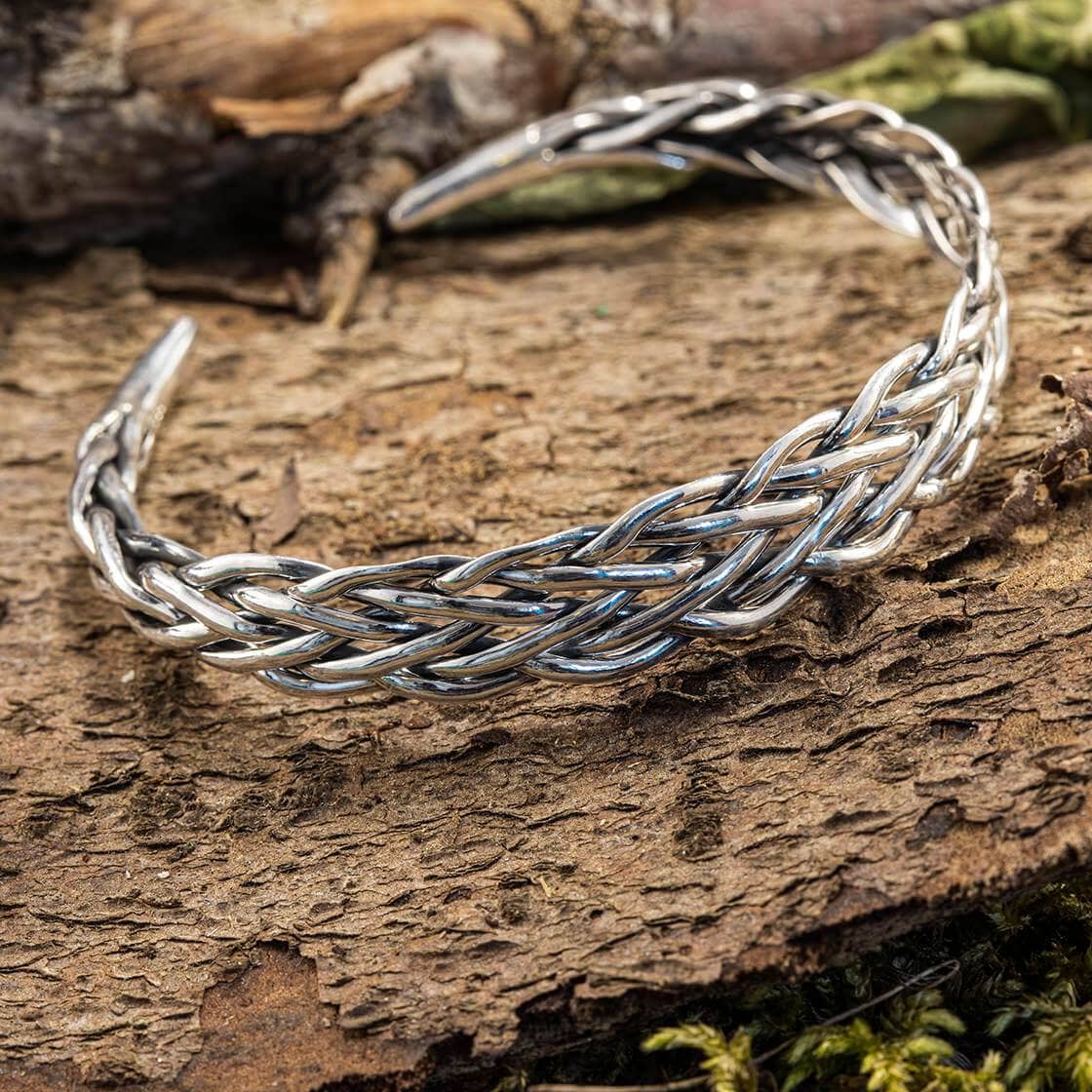 Et stykke robust, tekstureret bark fremhæver Armbånd Bangle Weave 925s Sterling Sølv. Det indviklede design viser sammenvævede tråde, der skaber et symmetrisk mønster. Antydninger af mos og løv i baggrunden understreger et naturligt udendørsmiljø.