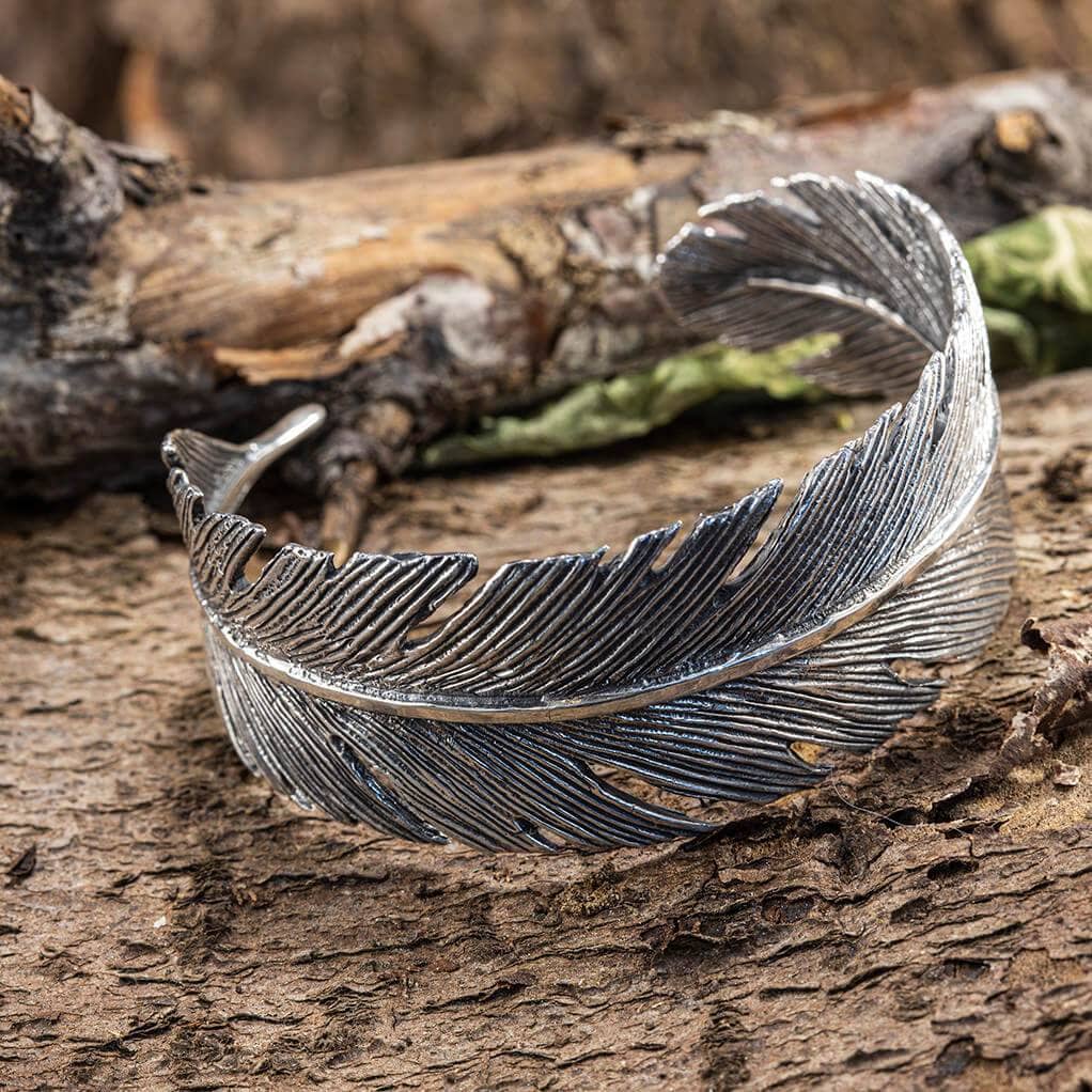 Et indviklet designet Armbånd Bangle Feather 925s Sterling Sølv, udformet til at ligne en fjer, ligger elegant på en tekstureret træoverflade med en blødt sløret baggrund. Den omhyggelige opmærksomhed på detaljer i fjerens tekstur sammen med den naturlige indstilling forstærker den organiske følelse af dette fortryllende armbånd.