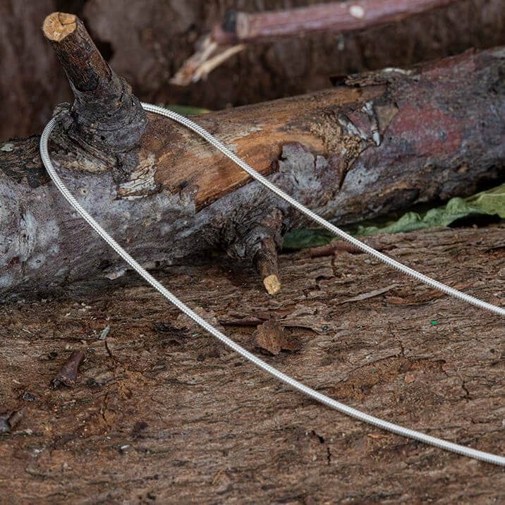 Halskæde Slangekæde 925s Sølv 1mm er draperet over en ru trægren, der ligger på en skovbund. Baggrunden inkluderer bark, snavs og nedfaldne blade, hvilket bidrager til en naturlig, robust indstilling.