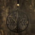 Load image into Gallery viewer, En Mobil Yggdrasil Wood Viking, et sort cirkulært ornament prydet med et symmetrisk trædesign, der minder om Yggdrasil fra Nordisk mytologi, hænger mod en sløret mørk baggrund. Ornamentet er ophængt af en sort snor toppet med en lille gylden perle.
