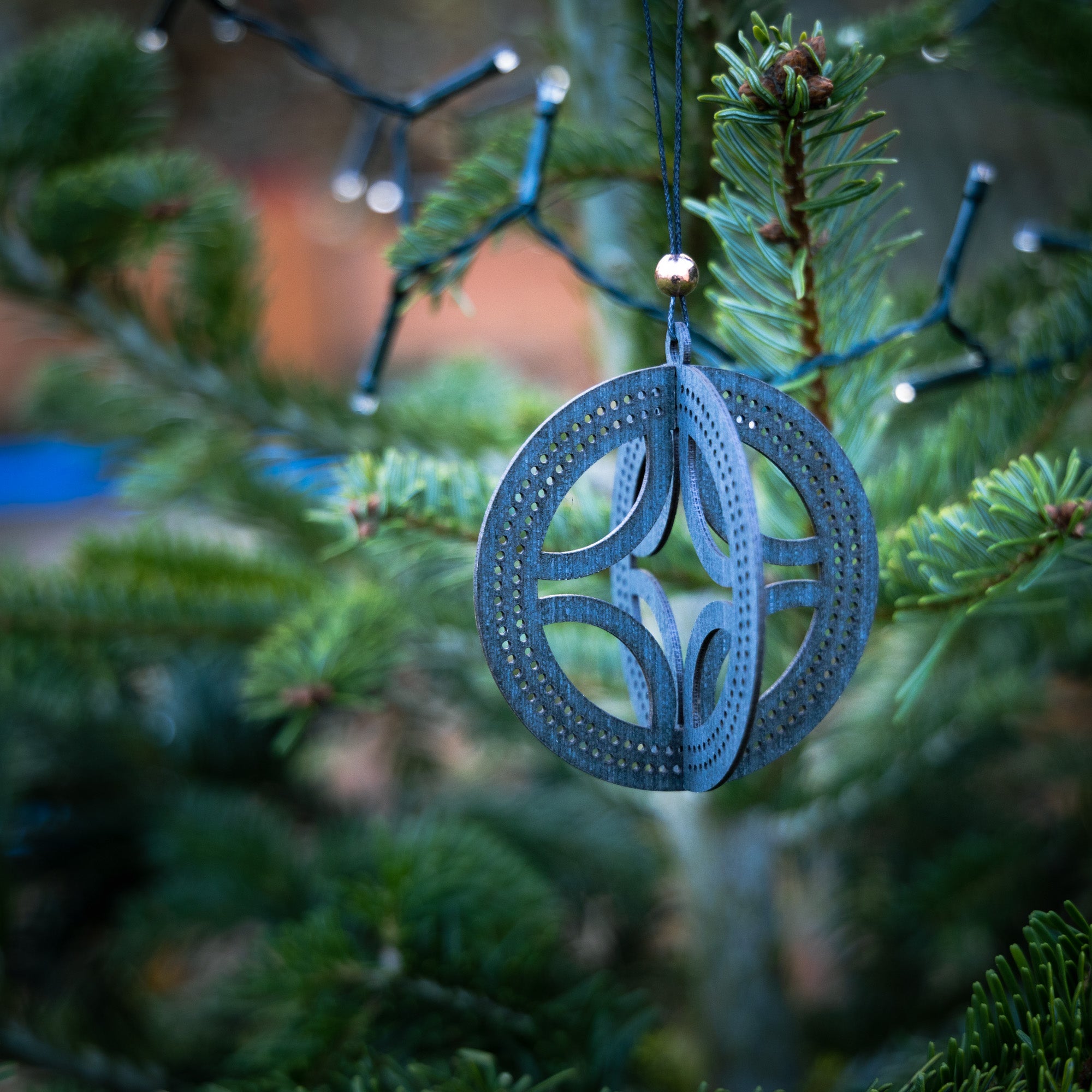 Et nærbillede af Mobil Solhjulet Wood Viking, et rundt, blåt ornament med et indviklet udskæringsmønster inspireret af oldnordisk kultur, hængende på en juletræsgren. Strengelys er delvist synlige i baggrunden blandt træets grønne nåle. Baggrunden er sløret, hvilket understreger det detaljerede håndværk i dette unikke stykke.