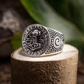 Load image into Gallery viewer, En Sølvring Thors Hammer 925s Sølv-ring med et detaljeret design hviler på en stenoverflade. Ringens ansigt viser et indviklet mønster med runesymboler, der omkranser et centralt billede af den nordiske hammer, Mjolnir. Siderne har hvirvlende og prikkede designs, mens grønne blade sløres i baggrunden.
