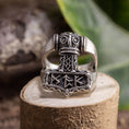 Load image into Gallery viewer, Sølvring Thors Hammer 925s Sølv, en ring i sterling sølv med indviklede nordiske designs, sidder på en træsokkel. Thors-hammeren fungerer som midtpunktet, prydet med runer og detaljerede udskæringer. Dette udsøgte stykke, fås i forskellige ringstørrelser, er sat mod grønt løv og en sløret naturlig indstilling.
