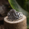 Load image into Gallery viewer, En detaljeret Sølvring Thors Hammer 925s Sølv, der viser et indviklet vikinge-inspireret design, sidder på en træoverflade. 925s Sterling sølv-ringen har udsmykkede mønstre, der ligner Thors hammer og er omgivet af grønt i den slørede baggrund.
