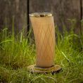 Load image into Gallery viewer, Et "Drikkeglas Horn med mønster" (300ml) med guldkant og diamantformede graveringer, fyldt med en brun væske, står på en træskranke midt i højt grønt græs med en sløret træbaggrund.
