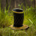 Load image into Gallery viewer, Et Drikkeglas Horn med Mønster (100 ml), med et indviklet sort design med guldaccenter, er placeret på en træskranke omgivet af levende grønt græs. Baggrunden er let sløret, hvilket gør opmærksom på det unikke glass detaljerede mønster og skinnende finish.
