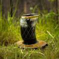 Load image into Gallery viewer, Et udskåret Drikkeglas Horn med Mønster (100ml) med en guldkantet kant sidder på en træskive midt i frodigt grønt græs. Drikkehornet fremviser indviklede geometriske mønstre og kan prale af en poleret, skinnende overflade. Baggrunden byder på sløret grønt græs og et rustikt plankeværk.
