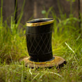 Load image into Gallery viewer, Et sort Drikkeglas Horn med Mønster (100 ml), indviklet designet med guldkant, hviler på et lille, rundt stykke træ i græsset. Baggrunden viser sløret grønt løv og en utydelig træstruktur, der fremkalder charmen og det unikke ved håndværksfremstillede stykker.
