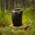 Load image into Gallery viewer, Et poleret Drikkeglas Horn med Mønster (100ml) med guldkant og indviklede udskårne mønstre er placeret på en træbund, omgivet af grønt græs. Baggrunden viser et rustikt plankeværk.
