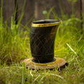 Load image into Gallery viewer, Et dekorativt Drikkeglas Horn med Mønster (100ml) med guldkant står på en træskive placeret på grønt græs. Skålen har et indviklet udskåret mønster og har en poleret finish, hvilket skaber et rustikt og naturligt udseende. Baggrunden er let sløret og fokuserer opmærksomheden på koppen.
