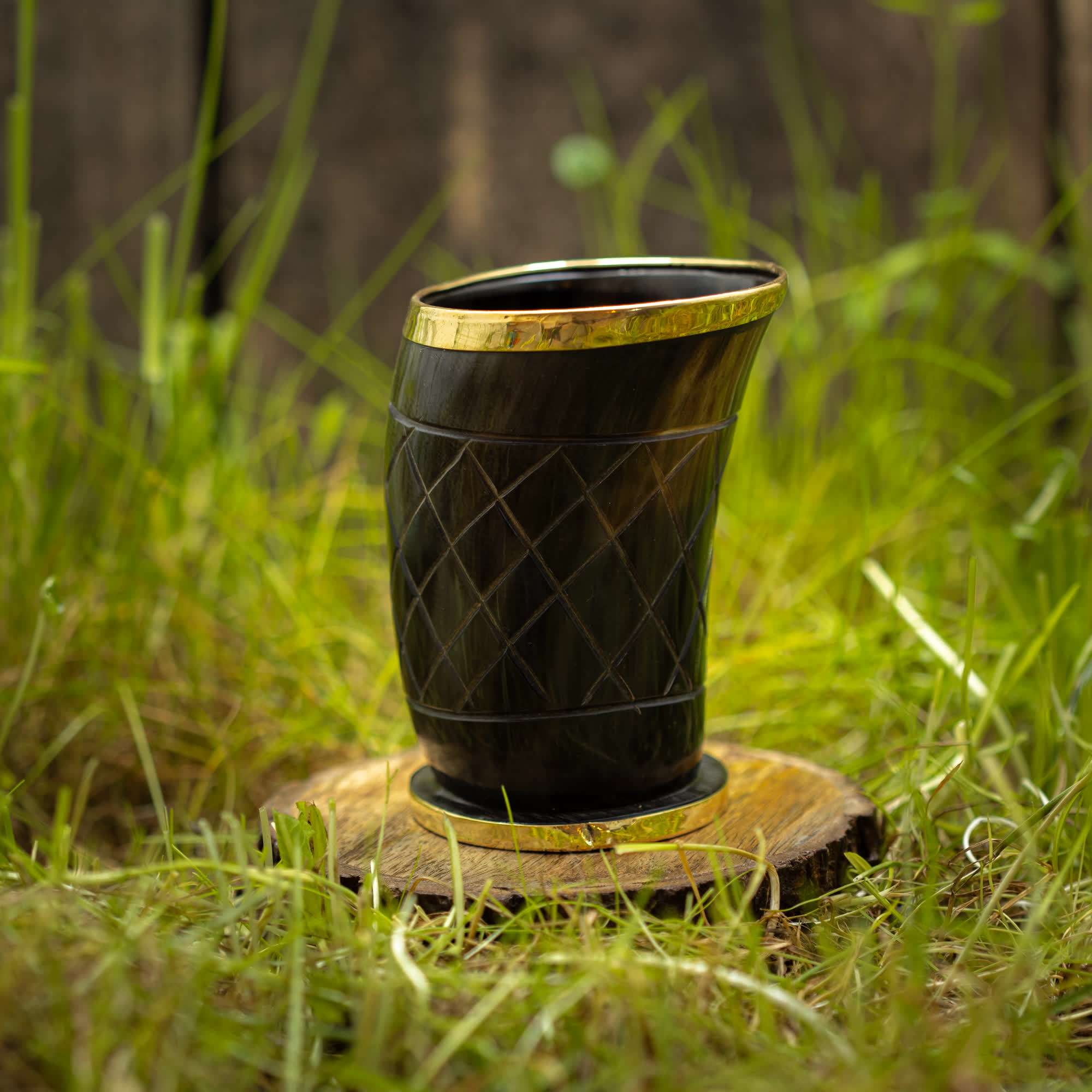 Et dekorativt Drikkeglas Horn med Mønster (100ml) med guldkant står på en træskive placeret på grønt græs. Skålen har et indviklet udskåret mønster og har en poleret finish, hvilket skaber et rustikt og naturligt udseende. Baggrunden er let sløret og fokuserer opmærksomheden på koppen.