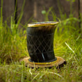 Load image into Gallery viewer, Et poleret, mørkt Drikkeglas Horn med Mønster (100ml) med en gylden kant sidder oven på en træbase på græs. Koppen har et krydsmønster ætset ind i overfladen og er sat mod en sløret baggrund med naturlige elementer.
