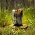 Load image into Gallery viewer, Et Drikkeglas Horn med Mønster (100ml) med en gylden kant sidder oprejst på en cirkulær træbund i græs. Hornet, der har et diamantmønster og en blank finish, er sat mod en baggrund af sløret grønt og træplanker, hvilket fremhæver dets unikke håndværk.
