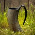 Load image into Gallery viewer, Vikingekrus Nature Horn (600-800 ml), et rustikt, buet drikkehorn med en poleret finish og fremtrædende håndtag, står på en plet af grønt græs. Træbaggrunden og de naturlige omgivelser forstærker dens middelalderlige, historiske tiltrækningskraft, der minder om den ældgamle vikingekrus, der blev brugt i oldtiden.
