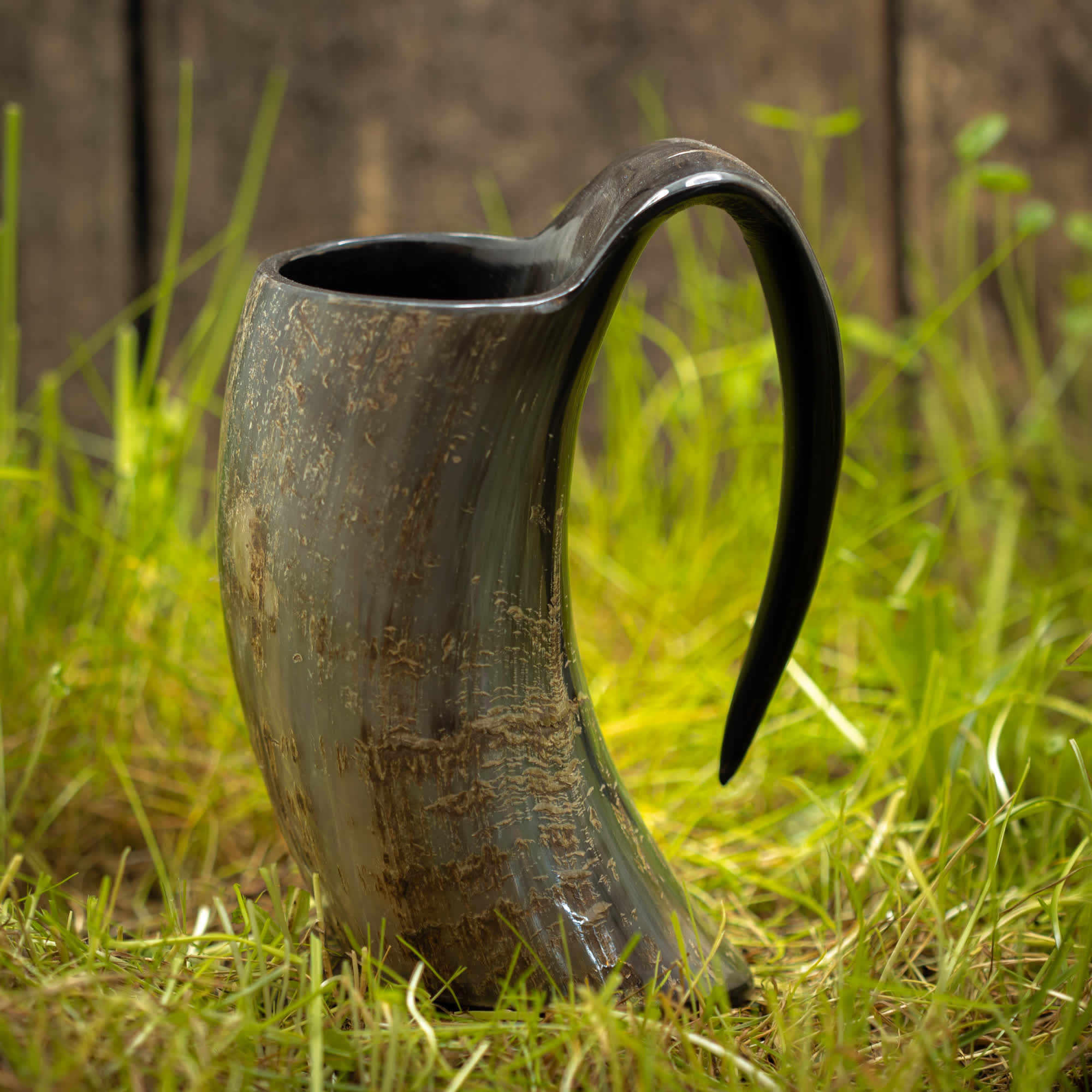 Vikingekrus Nature Horn (600-800 ml), et rustikt, buet drikkehorn med en poleret finish og fremtrædende håndtag, står på en plet af grønt græs. Træbaggrunden og de naturlige omgivelser forstærker dens middelalderlige, historiske tiltrækningskraft, der minder om den ældgamle vikingekrus, der blev brugt i oldtiden.