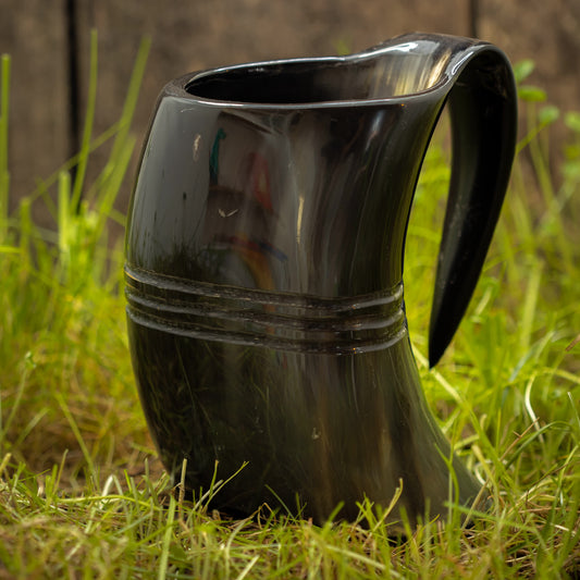 En Vikingekrus Helleristning Horn (600-800ml) kande med et håndtag hviler på græsset udendørs. Den sorte hornformede kande, der minder om store drikkekrus, har to vandrette riller i sin midterdel, hvilket tilføjer dets unikke design. En træoverflade er sløret i baggrunden.