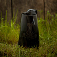 Load image into Gallery viewer, Et sort keramik Vikingekrus Thors Hammer Horn (600-800ml) med et håndtag og en tud, med et stiliseret Thors hammerdesign ætset på forsiden, er placeret på en plet af grønt græs mod en træbaggrund. Denne vikingekrus udstråler robust charme.

