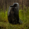 Load image into Gallery viewer, Et Vikingekrus Thors Hammer Horn (600-800 ml) med indviklede udskæringer, der ligner Thors hammer, sidder på grønt græs. Dette sorte drikkehorn står stolt på baggrund af forvitrede træplanker og tilføjer et rustikt præg til scenen.
